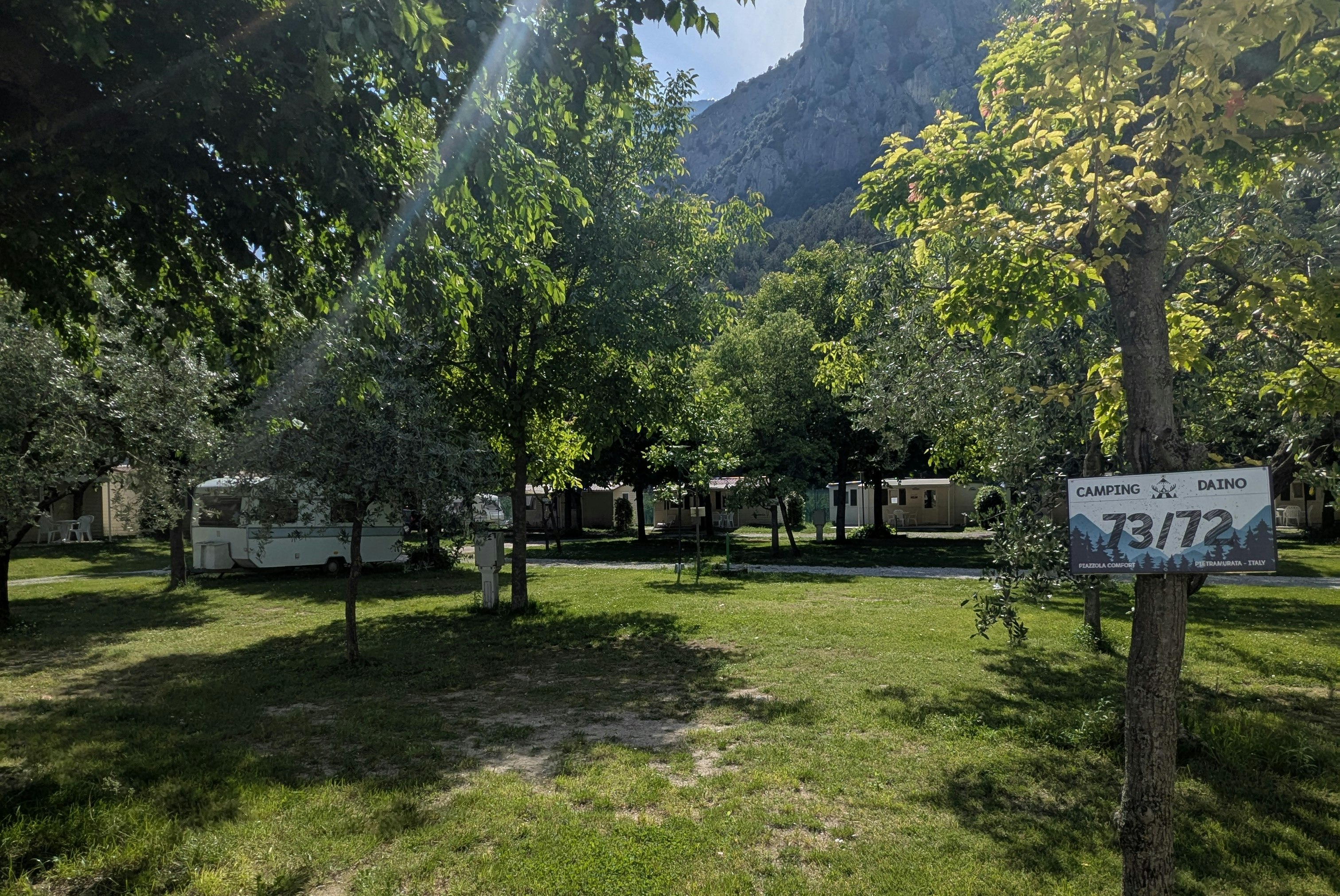 Campingplatz mit Wohnwagen und Bungalows, Bäume im Vordergrund und Blick auf die Berge, Schild „Camping Daino“.