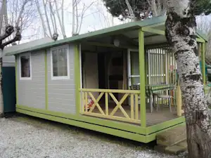 Grüner Wohnwagen mit Terrasse, Holzgeländer und Tisch, daneben Bäume und Kiesweg am Campingplatz.
