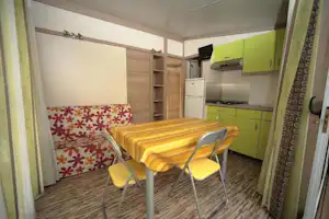 Innenansicht eines Camping-Mobilheims mit Esstisch, Stühlen und grüner Küchenzeile, Holzregal und Vorhängen am Fenster.