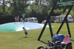 Spielplatz mit Hüpfburg auf Rasen, im Hintergrund Wohnwagen und Zelte zwischen Bäumen.