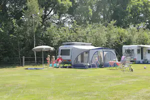 Wohnwagen mit Vorzelt und Markise auf dem Campingplatz, daneben Gartenmöbel und Fahrräder, umgeben von Bäumen.