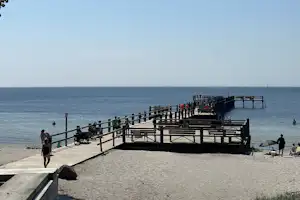 Holzsteg am Strand mit Geländer, Menschen gehen entlang, dahinter das Meer und ein kleiner Steg ins Wasser.