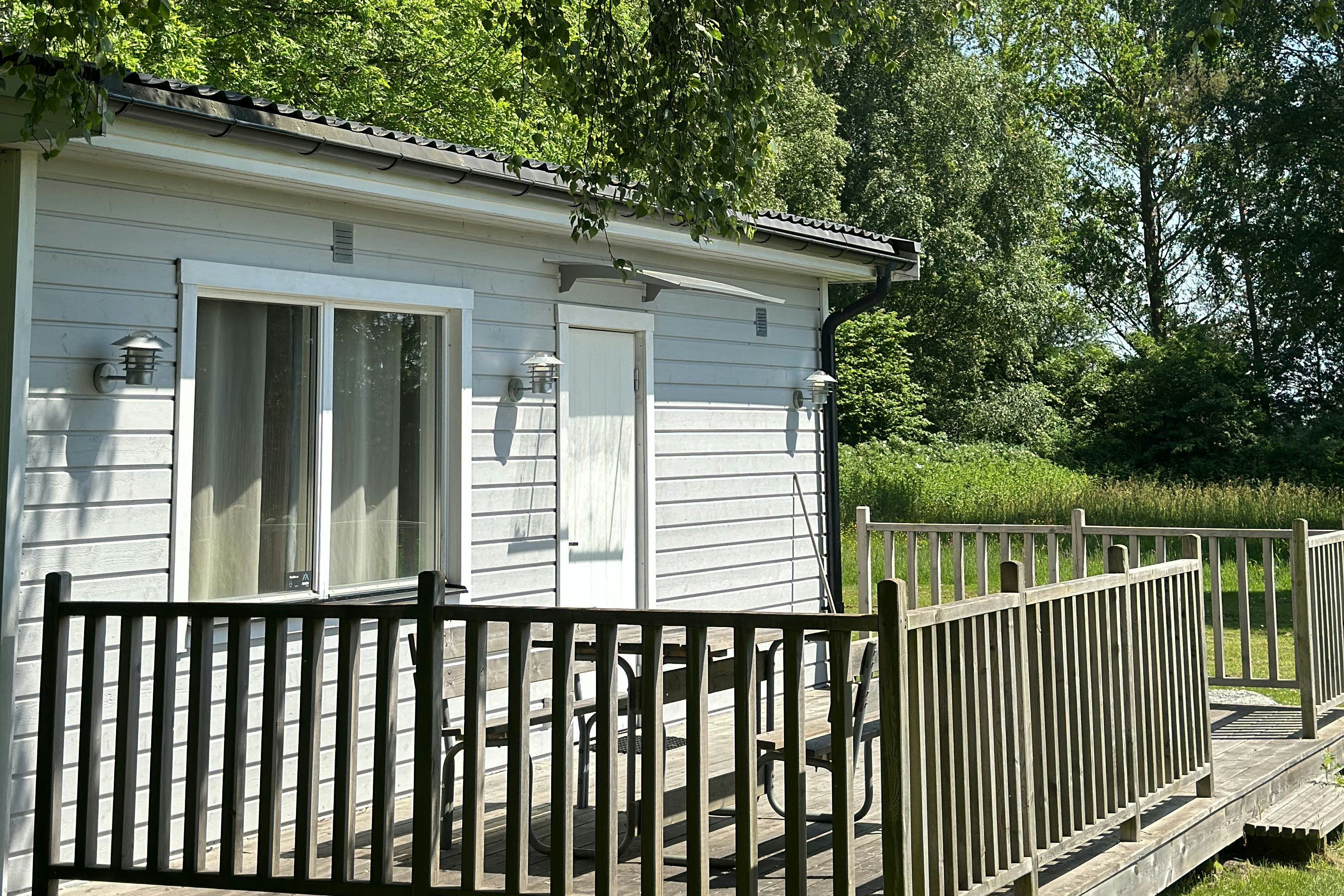 Bungalow mit Terrasse und Holzgeländer im Grünen, große Fensterfront, umgeben von Bäumen und dichtem Laub.