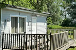 Bungalow mit Terrasse und Holzgeländer im Grünen, große Fensterfront, umgeben von Bäumen und dichtem Laub.