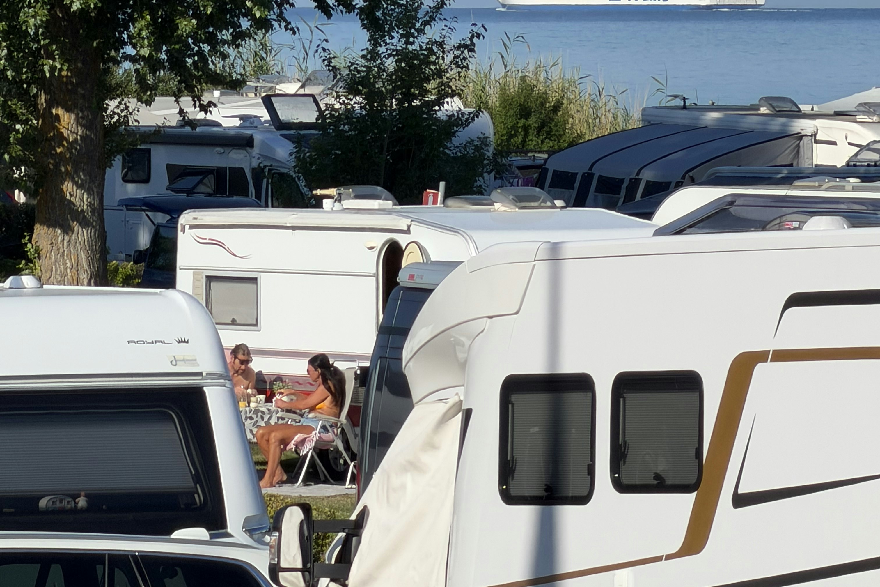Wohnmobile und Campingfahrzeuge stehen nebeneinander am Stellplatz, im Hintergrund ein Schiff auf dem Meer und Bäume am Rand.