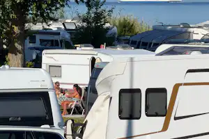 Wohnmobile und Campingfahrzeuge stehen nebeneinander am Stellplatz, im Hintergrund ein Schiff auf dem Meer und Bäume am Rand.