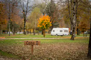 Wohnwagen auf einem Campingplatz im Herbst, im Vordergrund ein Schild „Cona D“ und Bäume mit gelben Blättern.
