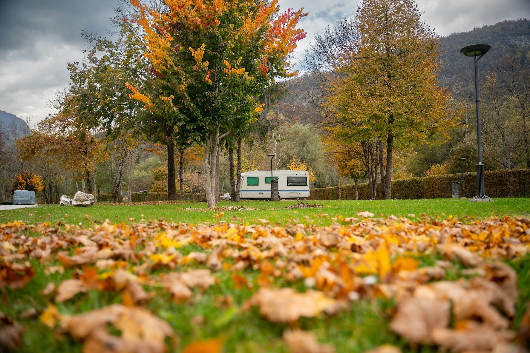Wohnwagen auf einem Campingplatz am grünen Rasen, im Vordergrund bunte Herbstblätter und Bäume mit Bergen im Hintergrund.