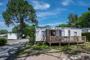 Mobilheim mit Holzveranda und Eingang, Kiesweg und Rasenfläche, umgeben von Bäumen und weiteren Wohnwagen auf dem Campingplatz.