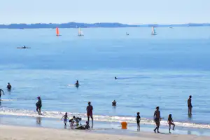 Badegäste am Strand am See, im Wasser Segelboote und Schwimmer, im Hintergrund ruhige Uferlinie.