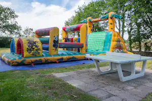 Bunte Hüpfburg auf einer Wiese im Campingbereich, daneben ein weißer Tisch und Bänke, im Hintergrund Bäume.