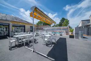 Terrasse mit vielen weißen Gartenstühlen und Sonnenschirm, daneben ein Pool und Gebäude mit Fenstern, im Hintergrund Bäume.