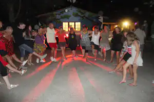 Gruppe von Campern tanzt nachts auf dem Weg vor einem beleuchteten Gebäude mit großen Fenstern und bunten Lichtern.