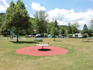 Campingplatz mit Rasenfläche, Feuerstelle und Stellplätzen mit Zelten, Wohnwagen und Autos zwischen Bäumen.