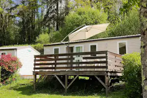 Mobilheim mit Terrasse und Sonnenschirm, Holzgeländer und Fensterfront, umgeben von Bäumen und grünem Garten.