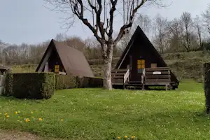 Zwei Camping-Häuschen mit Holzveranda und Treppe, umgeben von Hecken und grünem Rasen, im Hintergrund Bäume.