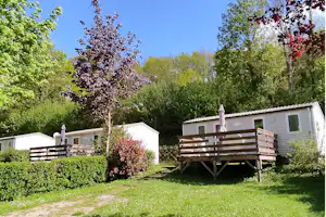 Wohnwagen mit Holzterrasse, umgeben von Hecken und Bäumen, im Campinggelände mit weiteren Unterkünften im Hintergrund.
