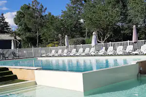 Camping-Pool mit türkisfarbenem Wasser, weißen Liegen hinter dem Zaun und Sonnenschirmen vor grünen Bäumen.