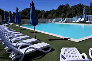 Camping-Pool mit türkisfarbenem Wasser, weißen Liegen und blauen Sonnenschirmen auf Rasen, im Hintergrund Zaun und Bäume.