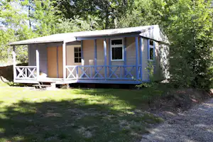 Mobilheim mit Veranda, Holzverkleidung und großen Fenstern, davor Sitzbank und Tisch auf Rasen im Grünen.
