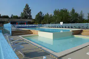 Schwimmbecken mit türkisfarbenem Wasser und Liegen auf dem Terrassenbereich, dahinter Bäume und ein Gebäude.