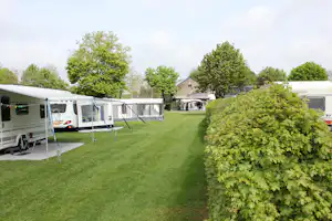 Reihen von Wohnwagen und Zelten auf einem gepflegten Campingplatz, mit Rasenweg und Bäumen im Hintergrund.
