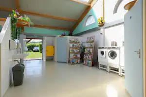 Innenraum eines Campinghauses mit Küchenzeile, Regalen und Waschmaschine, daneben ein Wäschekorb und große Fensterfront.