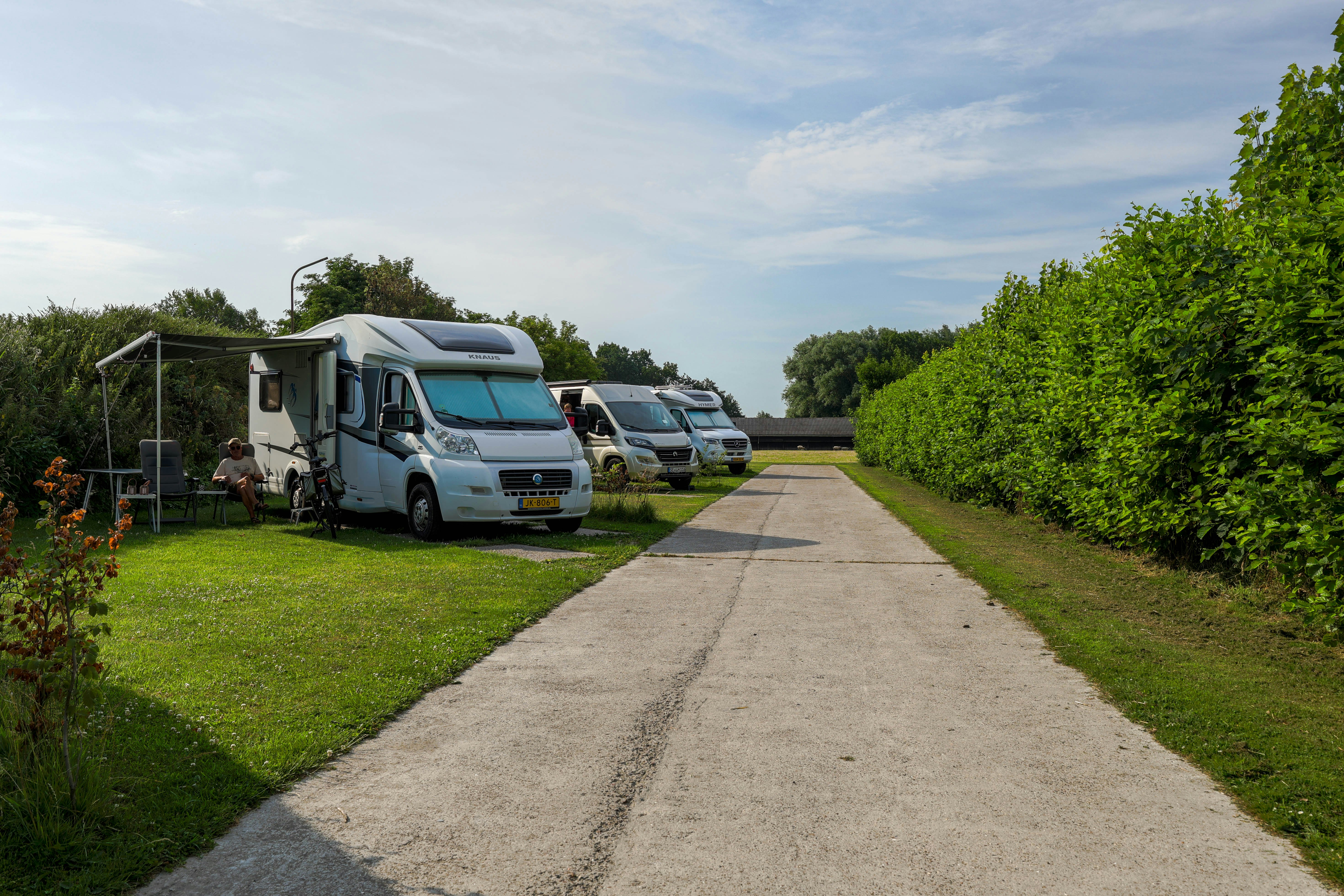 Wohnmobile auf einem Campingplatz entlang eines gepflasterten Wegs, mit Rasenflächen und Hecken, im Hintergrund weitere Fahrzeuge.