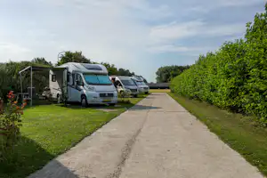 Wohnmobile auf einem Campingplatz entlang eines gepflasterten Wegs, mit Rasenflächen und Hecken, im Hintergrund weitere Fahrzeuge.