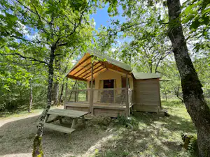 Holzhütte im Campingbereich mit Veranda und Sitzbank, umgeben von Bäumen und Kiesweg im Grünen.
