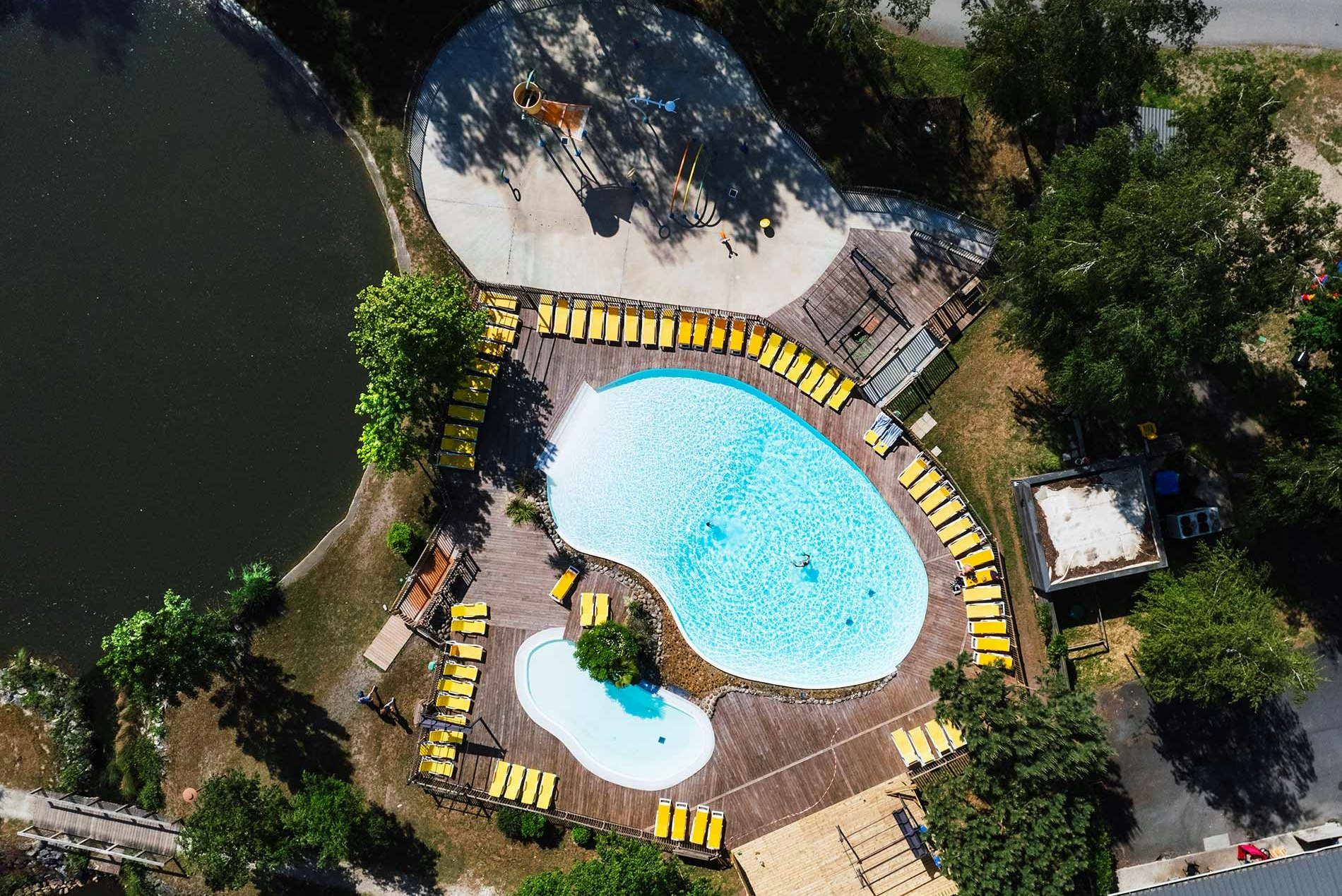 Großes blaues Schwimmbecken auf einem Campingplatz, umgeben von Bäumen, mit Holzwegen und Liegen am Rand.