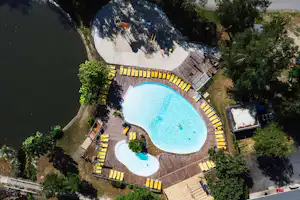Großes blaues Schwimmbecken auf einem Campingplatz, umgeben von Bäumen, mit Holzwegen und Liegen am Rand.