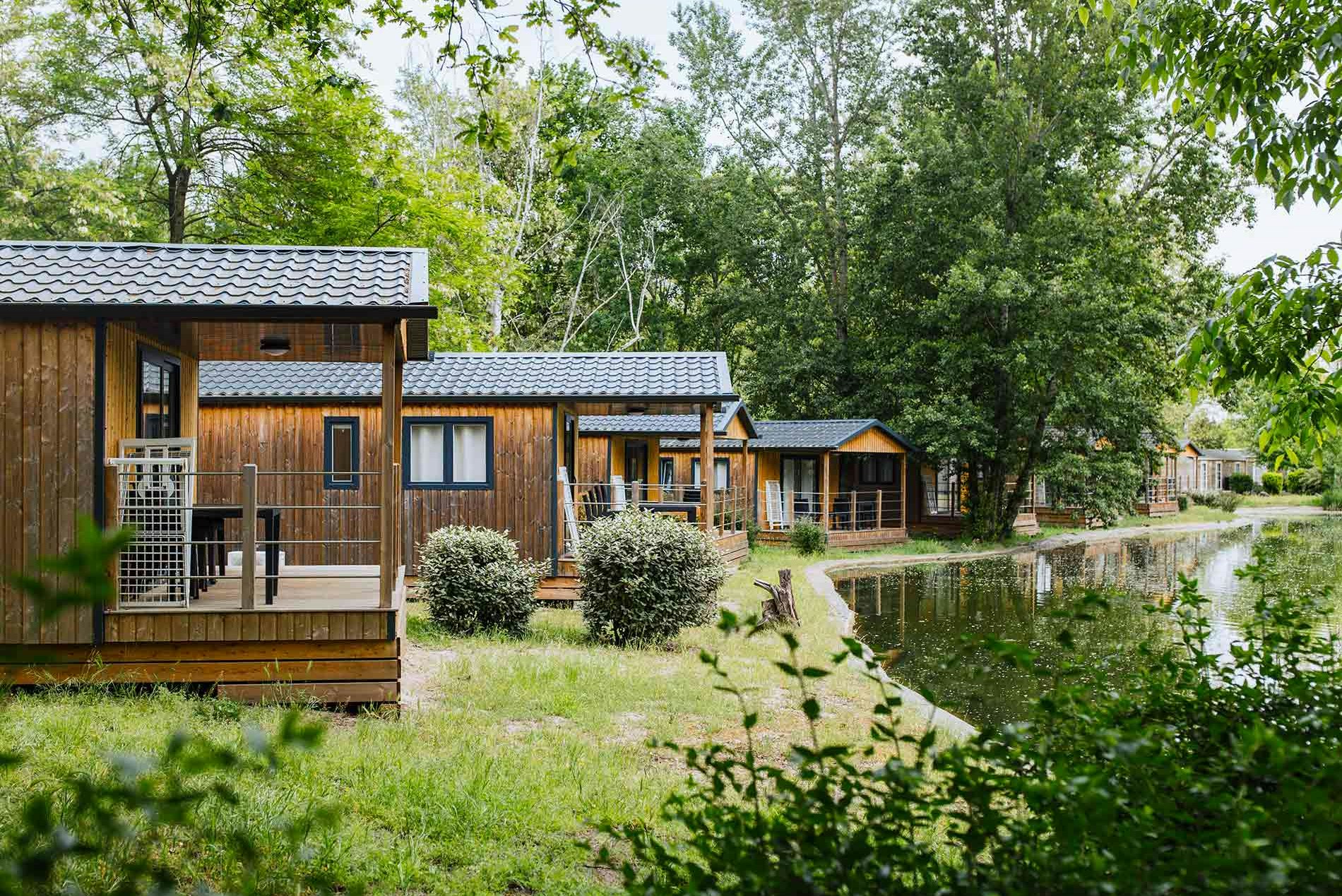 Holzhütten mit Terrassen und Sitzbereich, umgeben von Rasen und Büschen, im Hintergrund Bäume und ein Weg.