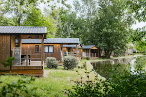 Holzhütten mit Terrassen und Sitzbereich, umgeben von Rasen und Büschen, im Hintergrund Bäume und ein Weg.