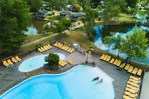 Schwimmbecken mit Liegestühlen am Campingplatz, daneben ein kleiner See und Bungalows zwischen Bäumen.