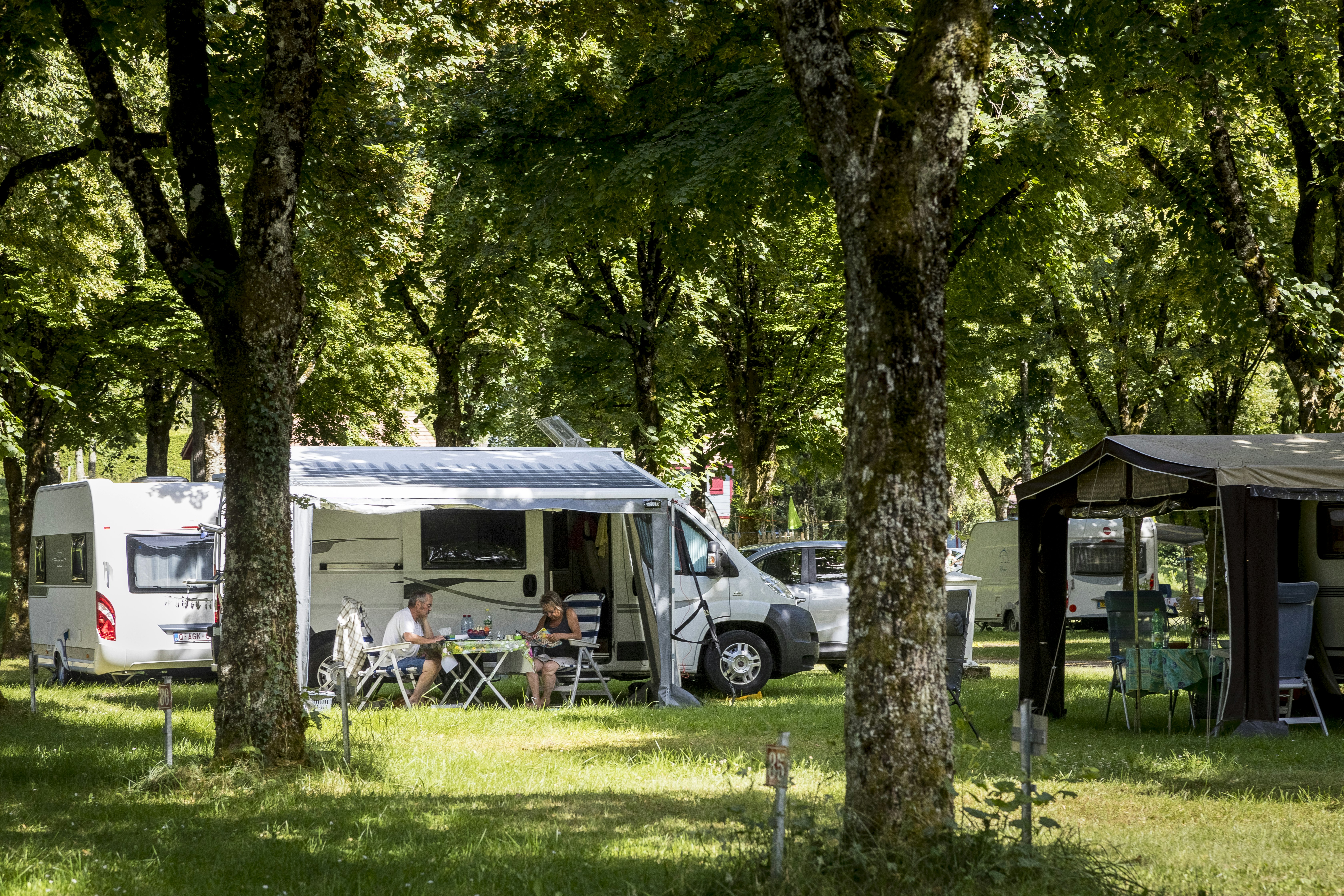 Campingplatz im Grünen: Wohnmobil mit Vorzelt, daneben Sitzgruppe und Tisch, im Hintergrund weitere Zelte zwischen Bäumen.