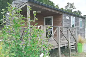 Holz-Bungalow mit Veranda und Geländer, davor Sträucher und ein grüner Eimer am Eingang.