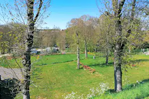 Grünes Campinggrundstück mit Rasenfläche, Bäumen und Hecken, im Hintergrund ein Gebäude und ein Weg.