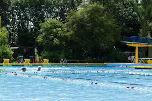 Schwimmbecken mit Bahnen und roten Markierungen, im Hintergrund Bäume und ein gelbes Sprungbrett am Campingplatz.