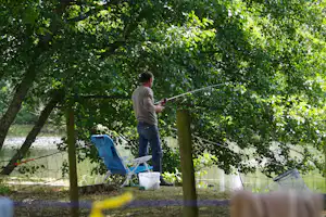 Angler steht am Flussufer zwischen Bäumen und hält eine Angelrute, daneben ein blauer Campingstuhl und Eimer.