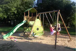 Grünes Spielzelt mit Rutsche und Schaukeln auf einem Sandspielplatz im Grünen, mit Kindern am Klettergerüst.