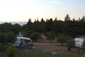 Campingplatz mit Wohnwagen und Vorzelten, Sitzgruppe und Grill auf dem Stellplatz, umgeben von Bäumen und Wiese.