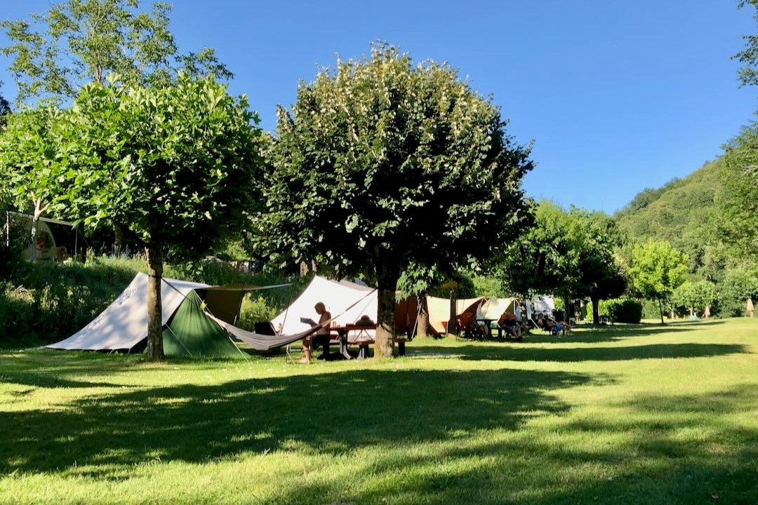 Zeltplatz mit mehreren Zelten auf einer grünen Wiese, Sitzbänke und Tische zwischen Bäumen vor einem grünen Hügel.