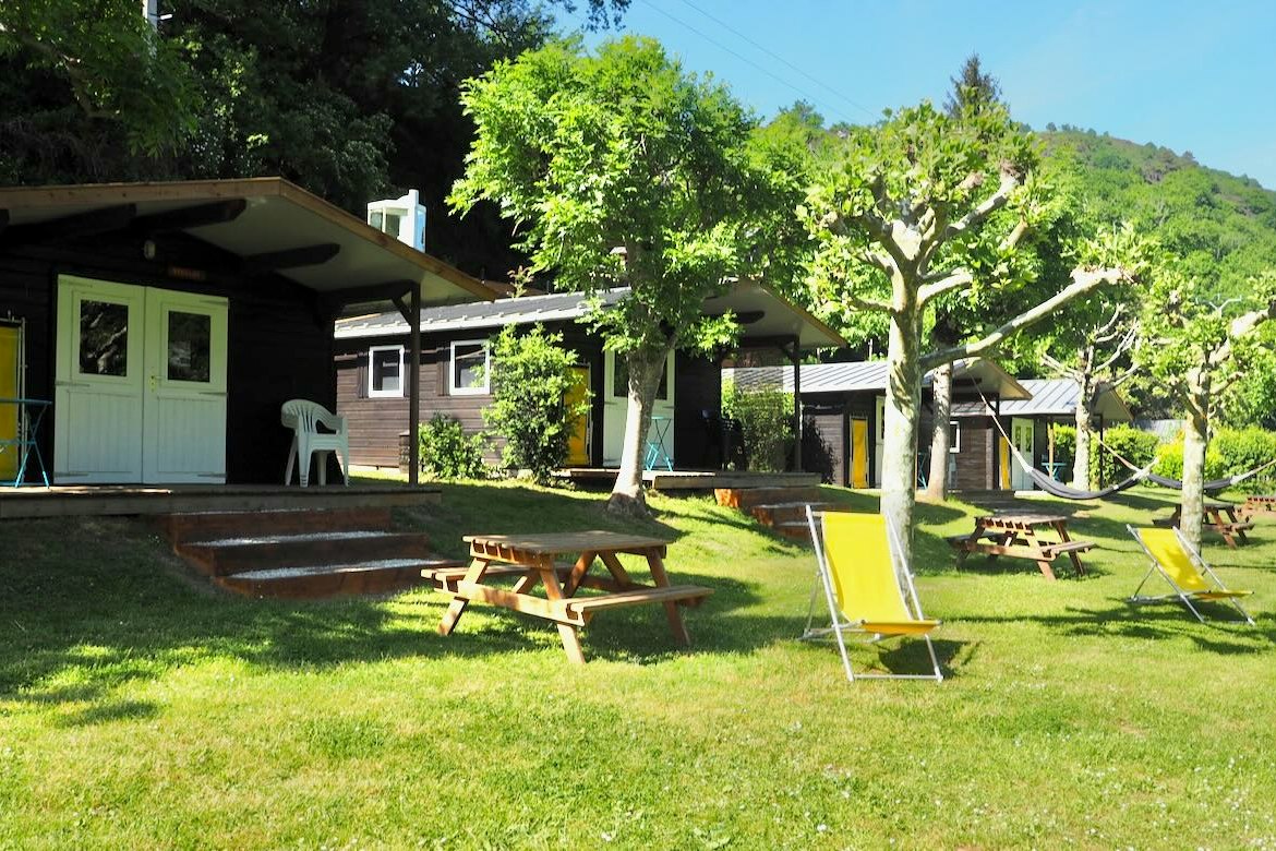 Campingplatz mit Mobilheim und Terrasse, Rasenfläche mit Picknicktisch und Liegestühlen, Bäume und Hügel im Hintergrund.