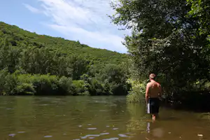 Mann steht im Wasser am Seeufer, umgeben von dichtem Grün und Bäumen, im Hintergrund bewaldete Hügel.