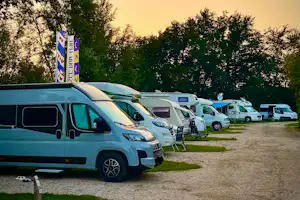 Reihe von Wohnmobilen und Campingbussen auf einem Kiesweg, davor ein Wohnmobil, im Hintergrund weitere Fahrzeuge und Bäume.
