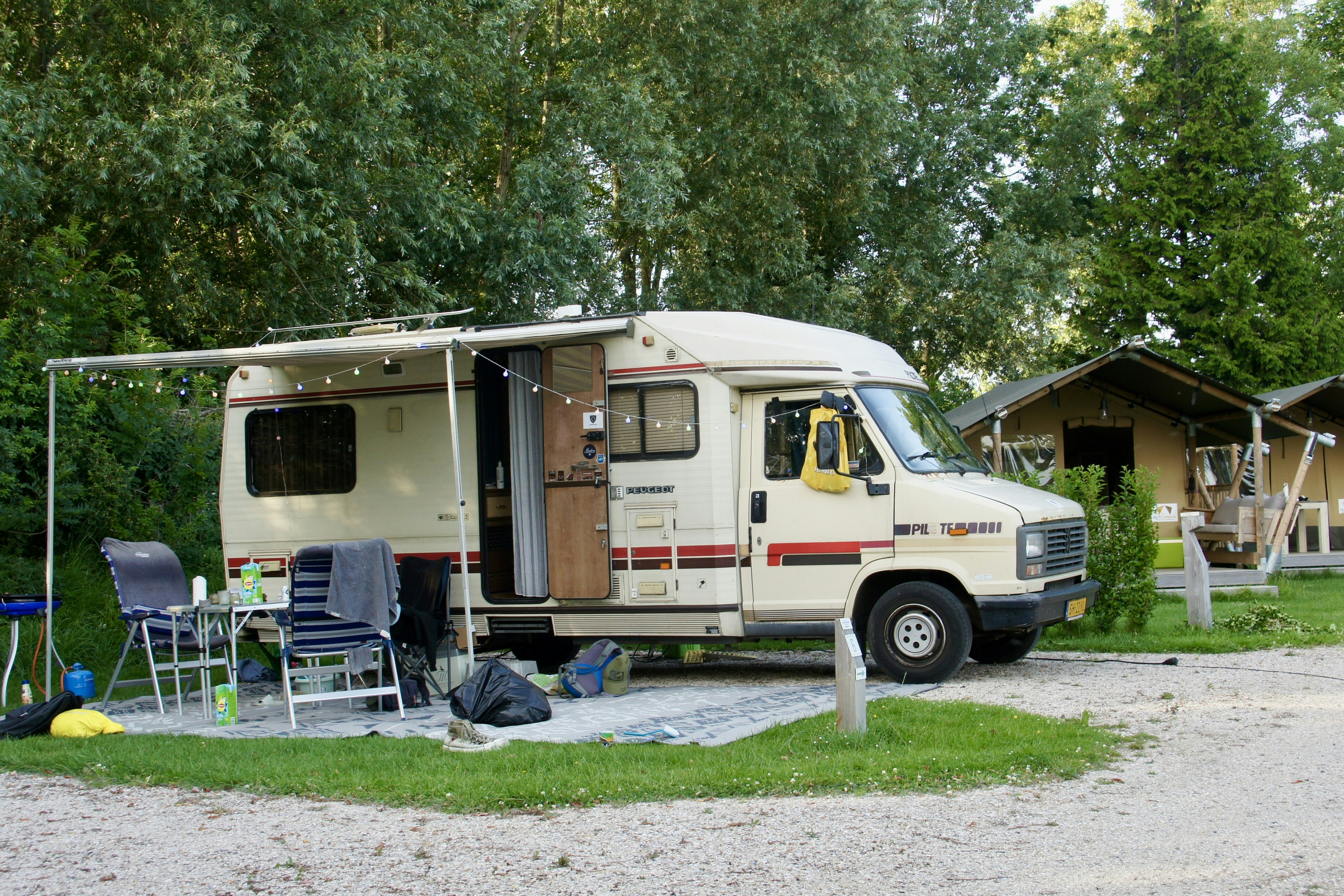 Wohnmobil am Stellplatz mit Vorzelt, Stühlen und Tisch auf dem Gras, im Hintergrund Bäume und weitere Campingunterkünfte.