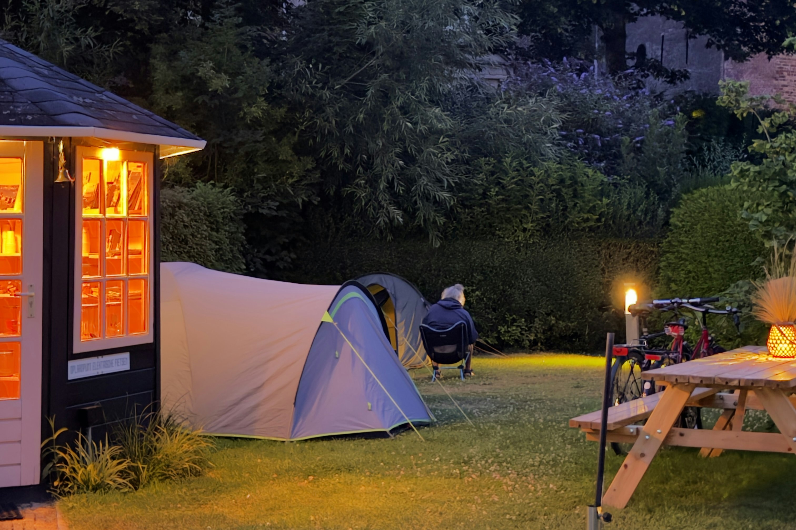 Zelt auf einem Campingplatz im Abendlicht, neben einem beleuchteten Sanitärgebäude und einem Holz-Tisch mit Lampe im Garten.