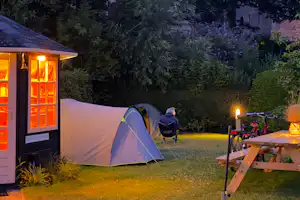 Zelt auf einem Campingplatz im Abendlicht, neben einem beleuchteten Sanitärgebäude und einem Holz-Tisch mit Lampe im Garten.