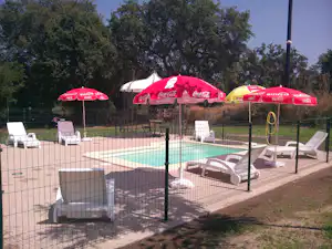Beheizter Camping-Pool mit Einfassung, Sonnenliegen und roten Sonnenschirmen im Garten, umgeben von Bäumen und Zaun.
